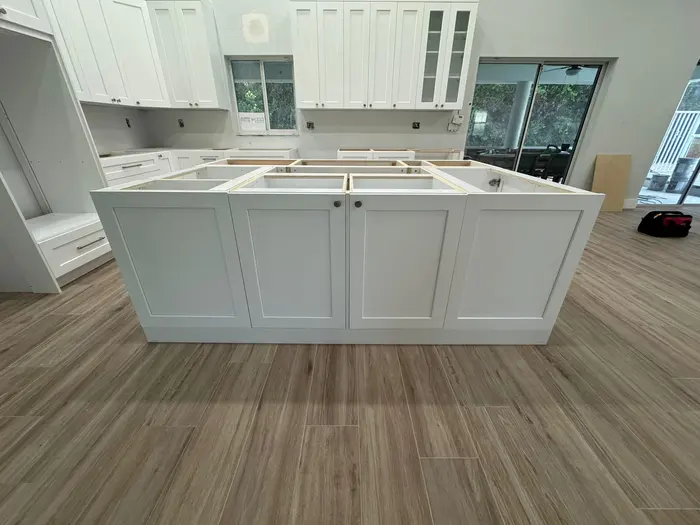 Mid-construction kitchen with white cabinet bases installed, wood-grain flooring laid, and upper cabinetry along walls with sliding glass doors to patio visible.