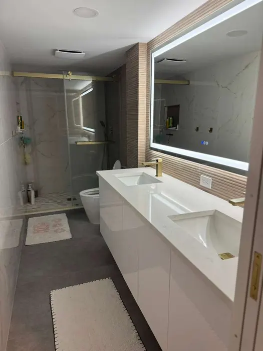 Modern finished bathroom with dual white vessel sinks, wood-accent vanity wall, LED-lit mirror, marble tile shower enclosure, and contemporary lighting fixtures