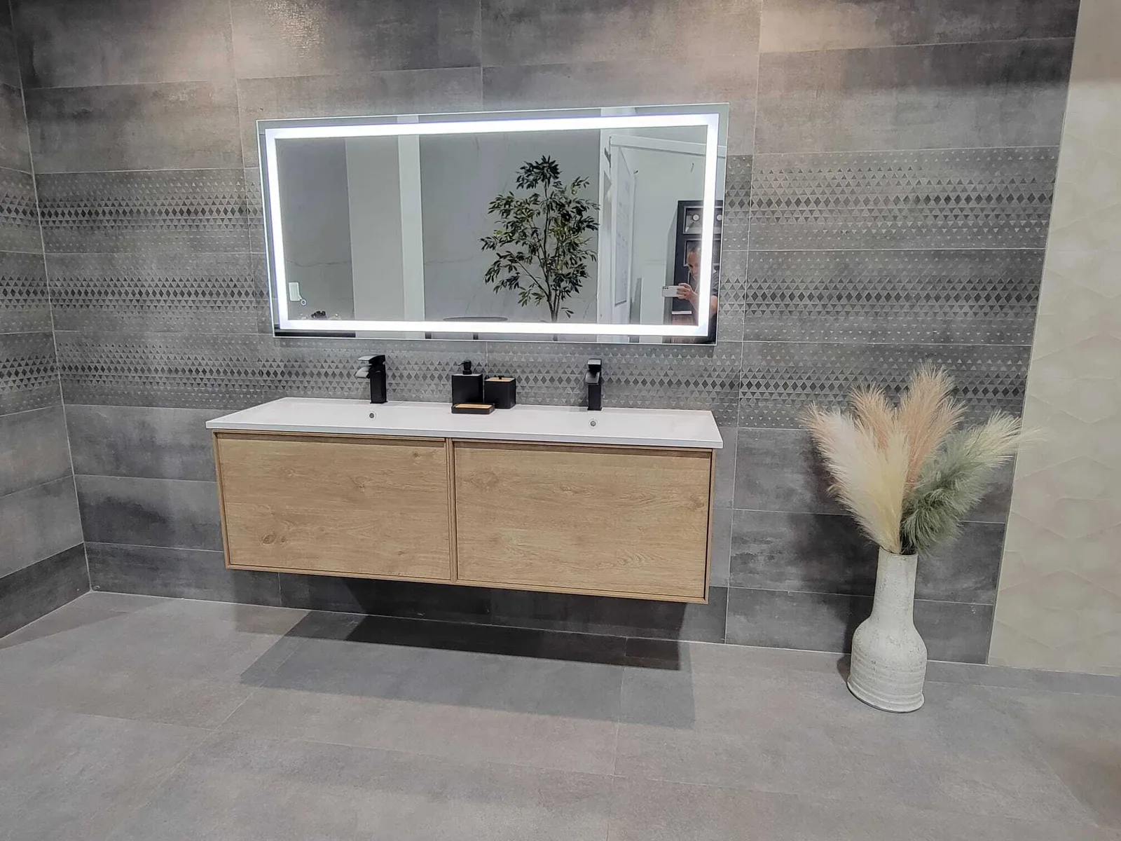 Modern completed bathroom vanity with floating natural wood cabinet, white double sink, LED-lit mirror, gray tile walls with geometric accent strip, and decorative accessories